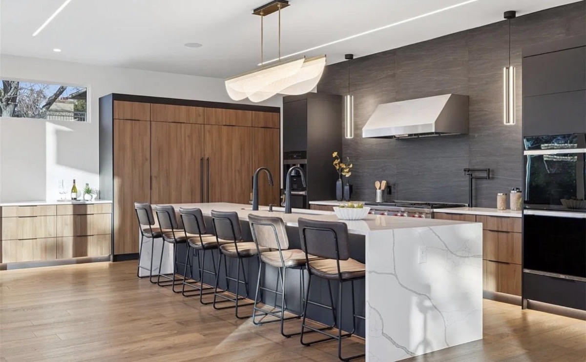 Modern kitchen with a waterfall quartz island, navy counter stools, walnut cabinets and dark tile wall