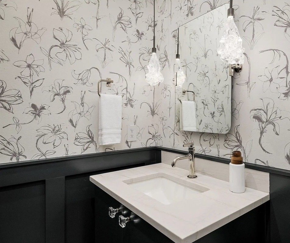 Powder room with black-and-white botanical wallpaper, black wainscoting, crystal pendant lights and a stone vanity