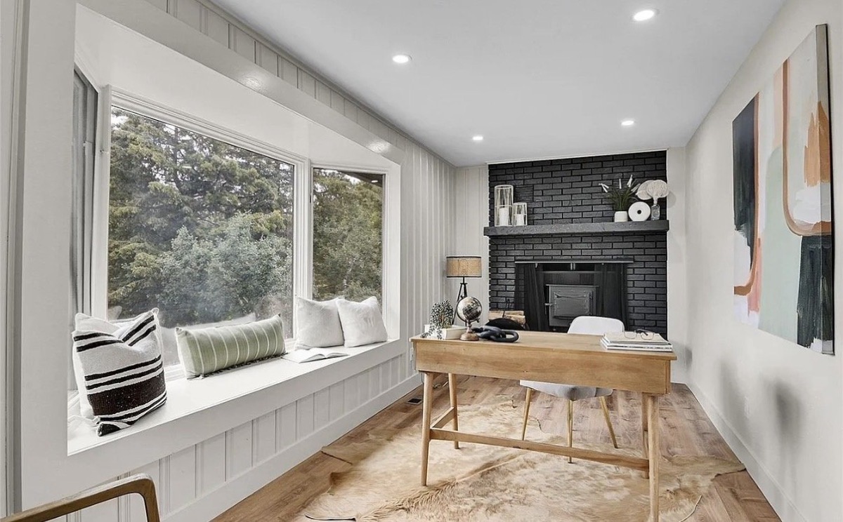 Sunroom office with a cushioned window seat, black painted brick fireplace and wood writing desk